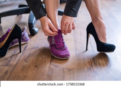 Eine Geschäftsfrau tauscht hohe Absätze gegen bequeme Schuhe am Arbeitsplatz aus. Eine Nahaufnahme der weiblichen Hände hebt ihre schwarzen Schuhe ab und setzt nach einem langen Arbeitstag farbige Turnschuhe im Büro an.