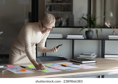 Business woman, female leader, executive, analyst working on project with documents, using online app on mobile phone, consulting internet, analyzing sales report, writing notes on paper stickers - Powered by Shutterstock