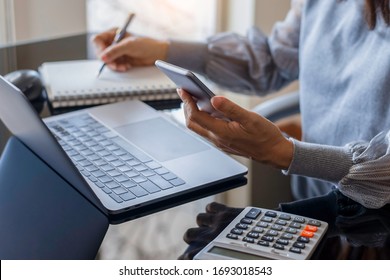 Business woman hand using mobile smartphone and writing on notebook, work on laptop computer with calculator on the desk at modern home office. Online working, work at home concept.