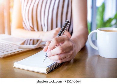 Business woman hand working at a computer and writing on a notepad with a pen in the office.On the wooden desk there is a cup of coffee and a mobile phone.