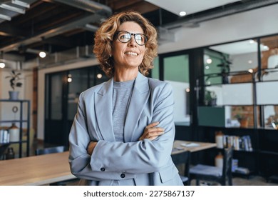 Business woman in her 40's looks away thoughtfully in an office. Professional woman standing in a suit with crossed arms. - Shutterstock ID 2275867137