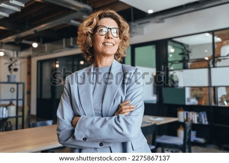 Business woman in her 40's looks away thoughtfully in an office. Professional woman standing in a suit with crossed arms.
