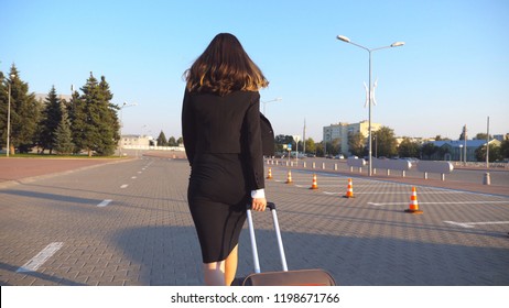 Business Woman With Her Suitcase Going From Airport To Taxi Parking. Lady Walking With Her Luggage Along City Street. Female Legs In High Heels Stepping At Sidewalk. Rear Back View Slow Motion.