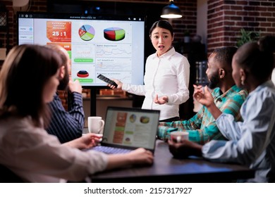 Business Woman Holding Remote Control Presenting Sales Pie Charts On Tv Screen Working Late In Night Meeting. Stressed Employee Doing Overtime With Team At Work Standing And Talking About Business