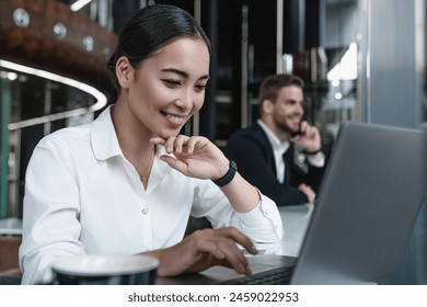 Business woman it-worker sitting at lounge cafe lobby coworking with laptop having coffee and looking on screen with male coworker on background. Freelancers working remote - Powered by Shutterstock