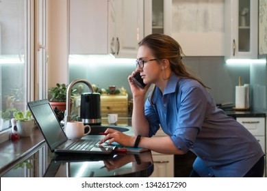 Business woman in kitchen at home. She is talking on cell phone and looking at laptop screen. Girl freelancer. Good morning.