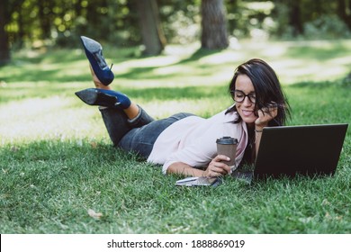 Business woman, lies in a summer grass park, using a laptop with a glass of coffee in hand. Remote work during quarantine. Woman with coffee. Outdoors. Soft selective focus.