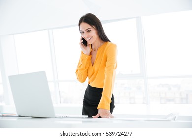 Business Woman In Orange Shirt Talking At Phone Near The Table And Looking At Laptop