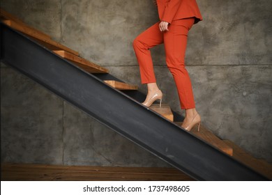 A Business Woman In An Orange Business Suit Climbs Up The Stairs. Career Ladder. High-heeled Shoes. Gray Background