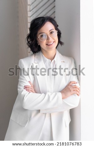 Business woman portrait. Middle-aged brunette in glasses and white suit folded her arms. Smiling at window. Looks into the camera.