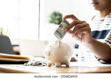 A Business Woman Is Putting Dollar Bills In A White Pig Piggy Bank To Save, She Is Doing An Income And Expense Account To Organize Her Financial Plan. Money Saving Ideas.