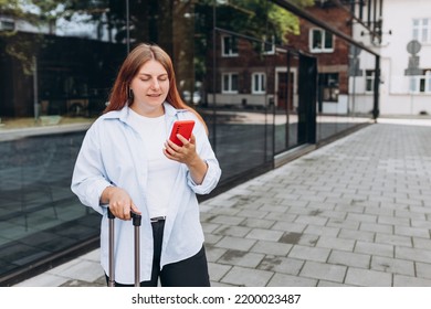 Business Woman With A Red Suitcase Using Smartphone In The European City. Hipster Girl Browsing Internet On A Phone And Waiting Taxi And Communicating Outdoors. Mobile Travel Maps.