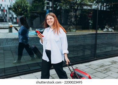 Business Woman With A Red Suitcase Using Smartphone In The European City. Hipster Girl Browsing Internet On A Phone And Waiting Taxi And Communicating Outdoors. Mobile Travel Maps.