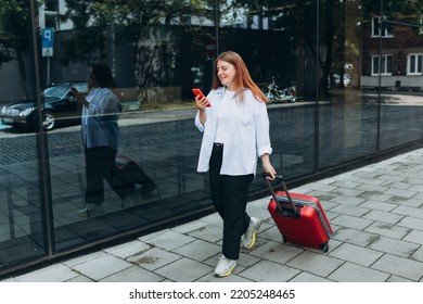 Business Woman With A Red Suitcase Using Smartphone In The European City. Hipster Girl Browsing Internet On A Phone And Waiting Taxi And Communicating Outdoors. Mobile Travel Maps.