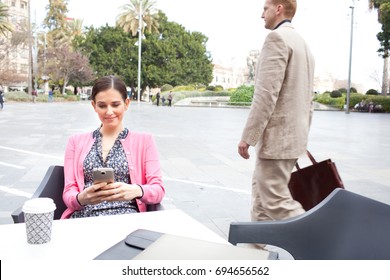 Business Woman Sitting At Coffee Shop Table Using A Smart Phone In City Exterior With Business Man Walking Passed Holding Briefcase, Financial Outdoors. Professional Female Using Technology, Smiling.