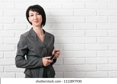 business woman with smartphone, dressed in a gray suit poses in front of a white wall