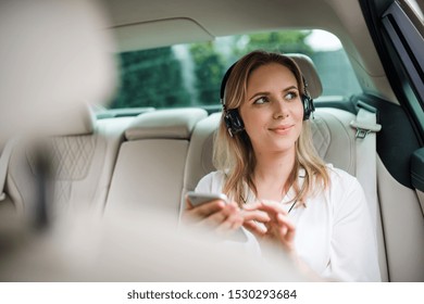 Business Woman With Smartphone And Headphones Sitting On Back Seats In Taxi Car.