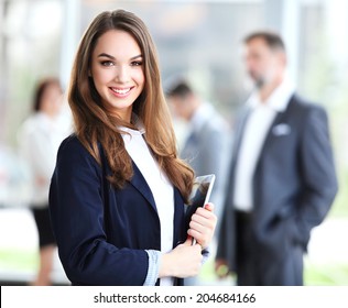 Business Woman Standing In Foreground With A Tablet In Her Hands, Her Co-workers Discussing Business Matters In The Background 