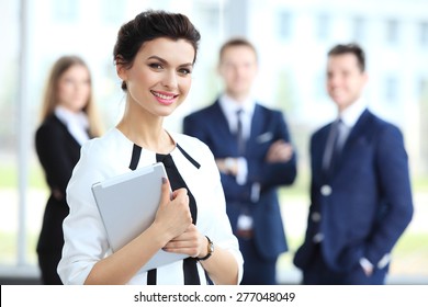 Business Woman Standing In Foreground With A Tablet In Her Hands, Her Co-workers Discussing Business Matters In The Background