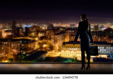 Business Woman Standing At The Window In The Night Office, Through The Window Illuminated City