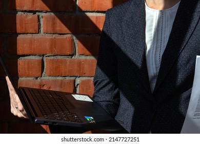 business woman in suit holding laptop and documents. Close-up Part of black jacket against on brick wall background with shadows, sunny day. Horizontal layout, selective focus.
