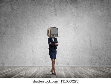 Business woman in suit with an old TV instead of head keeping arms crossed while standing inside empty room with gray wall on background.