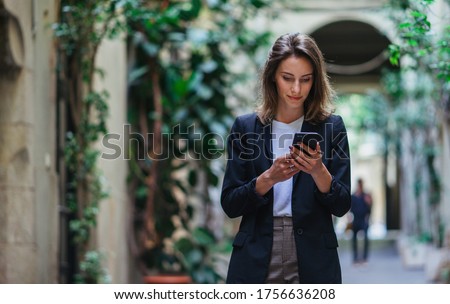 Business woman talking on mobile phone outdoors, professional woman in suit walking on old city street checks her smartphone
