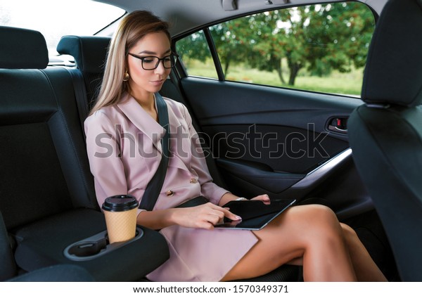 Business woman taxi car salon, summer day city, reads\
messages finger on touch screen activates application, tablet, work\
on road, secretary reads news. Casual makeup pink formal suit cup\
coffee tea
