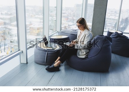 Business woman thinks while writing in a notebook at work. Modern successful woman. Young smiling girl in glasses sits in a chair at a table in an open office