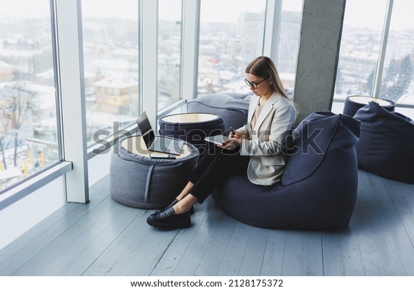 Business woman thinks while writing in a\
notebook at work. Modern successful woman. Young smiling girl in\
glasses sits in a chair at a table in an open\
office