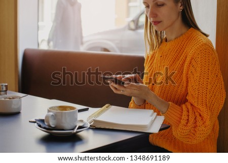 Business woman typing messages on smart phone in co-working, woman wearing orange sweater sitting in cafe