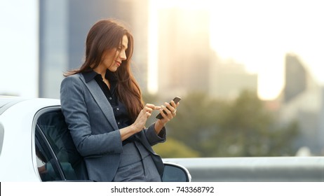 Business Woman Uses Smartphone While Leaning on Her Premium Class Car. Big City with Skyscrapers in the Background.