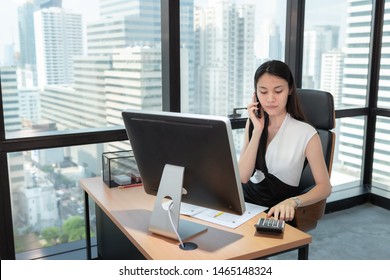 Business Woman Using Calculator And Talking On Mobile Phone In Her Office., Beautiful Of  Asian Woman Is Calling To Someone On Her Cell Phone While Calculating Data Saving At Her Table Desktop.