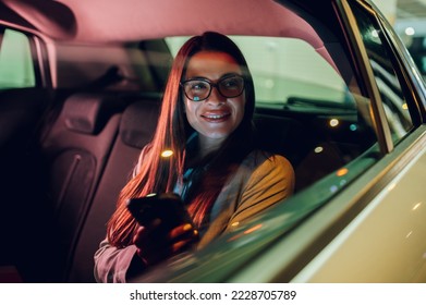 Business woman using smartphone while sitting in a backseat of a car at night. Beautiful female using mobile phone in a taxi in a city street with working neon signs. View trough the car window.