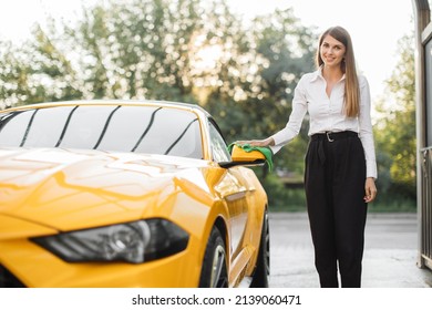 Business Woman Wearing On A White Shirt And Black Trousers Wiping Rearview Car Mirror Of Her Modern Luxury Car In A Self-service Car Wash Station Outdoors With A Green Microfiber Cloth