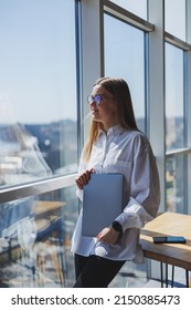 Business Woman In A White Shirt And Glasses With A Laptop In Her Hands In The Office By The Window. Modern Businesswoman In Stylish Clothes. Freelance Girl Working Outside The Office