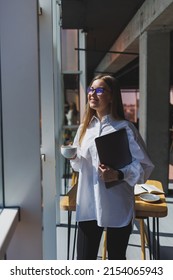 Business Woman In A White Shirt And Glasses With A Laptop In Her Hands In The Office By The Window. Modern Businesswoman In Stylish Clothes. Freelance Girl Working Outside The Office