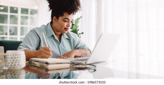 Business woman working from home writing notes while looking at laptop. Confident woman sitting at desk using laptop and taking notes.