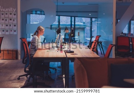business woman working overtime alone at his desk on a desktop computer in an open space modern coworking office