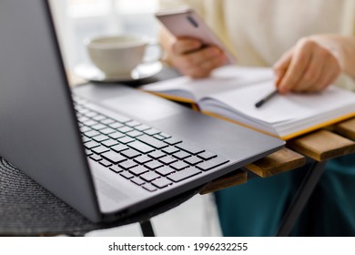 business woman works at laptop, holds smartphone in her hand and makes entries in diary. Business planning and tasks for day or week. Female in business. Keyboard laptop