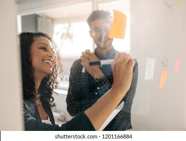 Business Woman Writing On Sticky Notes Pasted On Transparent Glass Wall In Office. Office Colleagues Discussing Business Ideas And Plans On A Transparent Glass Board.