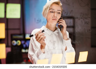 Business Woman Writing On Transparent Glass Wall With Sticky Notes In Office. Confident Focused Blond Lady, Discussing Business Ideas And Plans Talking On The Phone With Colleague