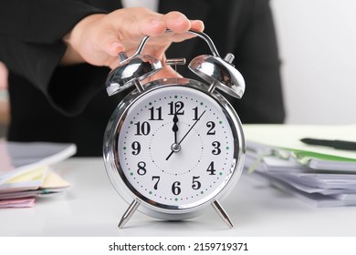 Business Woman's Hand Reaches Out To Touch Alarm Clock To Stop Time While Sitting At Her Office Desk. Selective Focus Of Alarm Clock On Table, Hands On Twelve O'clock. Concept Of Time Management