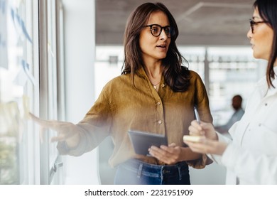 Business women discussing their ideas using sticky notes in an office. Female business professionals standing next to a window and brainstorming. - Powered by Shutterstock