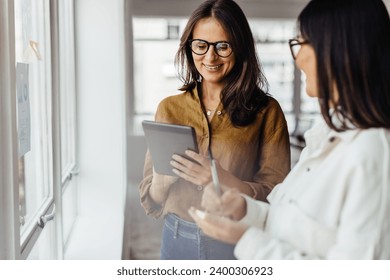 Business women standing next to a window and discussing their ideas. Female business professionals brainstorming using stocky notes and a tablet.