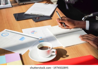 Business And Work Place Concept. Closeup Of Black Businessman Hand Holding A Pen On Working Desk With Document Notbook, Cup Of Coffee, Smartphone And Tablet