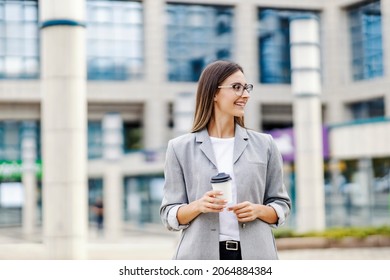 Business Young Woman And Coffee To Go. Successful Woman In Casual Clothes Stands In Front Of Business And Holding Coffee To Take Away In Her Hands While Looking To The Side With A Smile