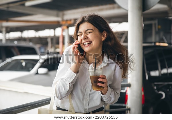 Business young woman with coffee talking\
cheerfully on the phone in the parking\
lot.