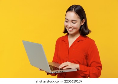 Businesslike Smiling Young Woman Of Asian Ethnicity 20s Years Old Wears Orange Shirt Hold Use Work On Laptop Pc Computer Typing Searching Chatting Isolated On Plain Yellow Background Studio Portrait