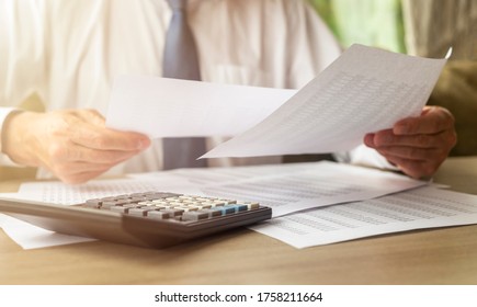 Businessman Analyzing Accounting Financial Documents With Calculator On Wooden Desk In Office. Man Holding Documents Over The Table And Compare Or Read Documents